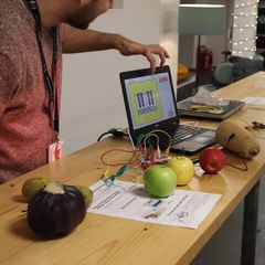 A volunteer pointing at a laptop that is connected to different pieces of fruit using some wires
